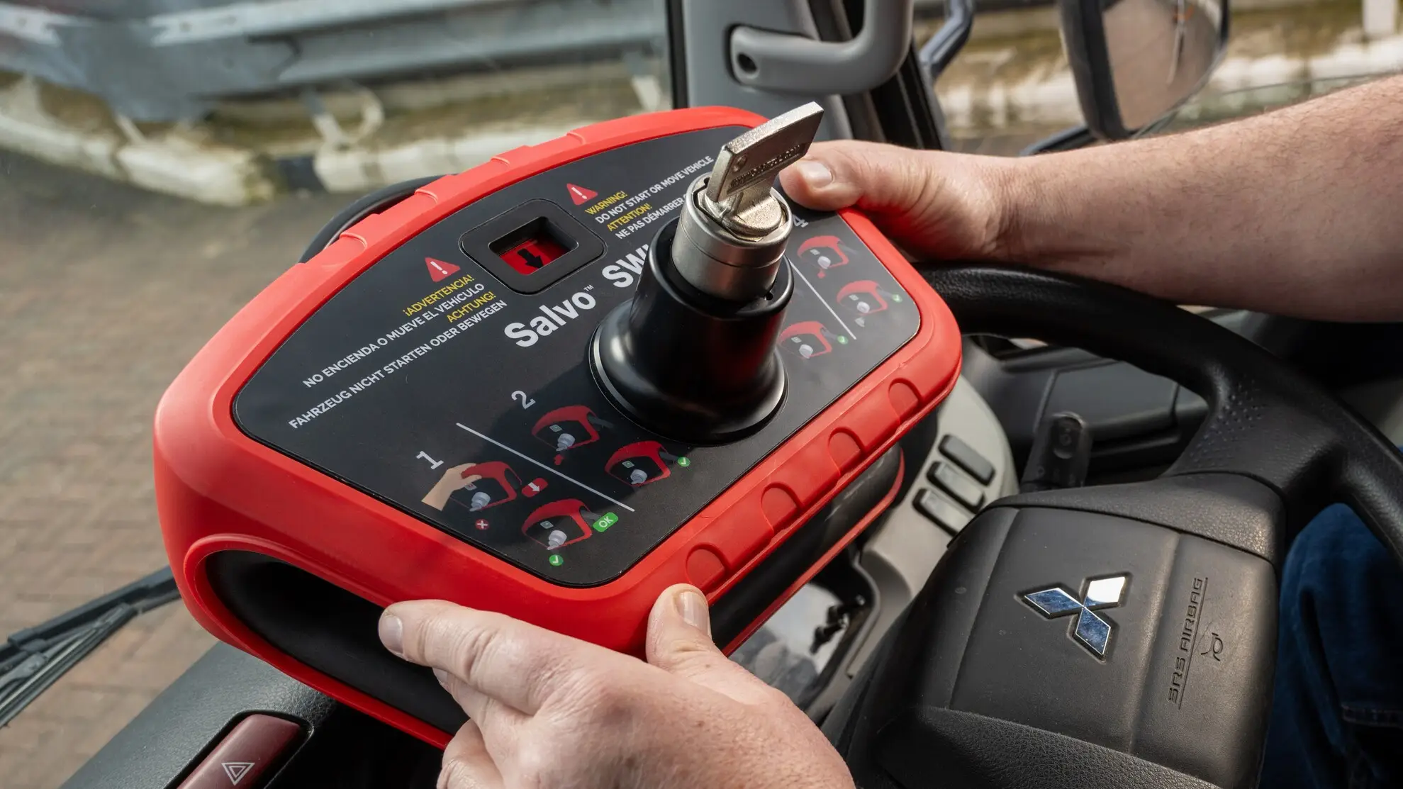 Driver inserting the key into a Salvo Steering Wheel Lock (SWL) mounted on a vehicle's steering wheel, with multilingual warning labels not to start or move the vehicle.