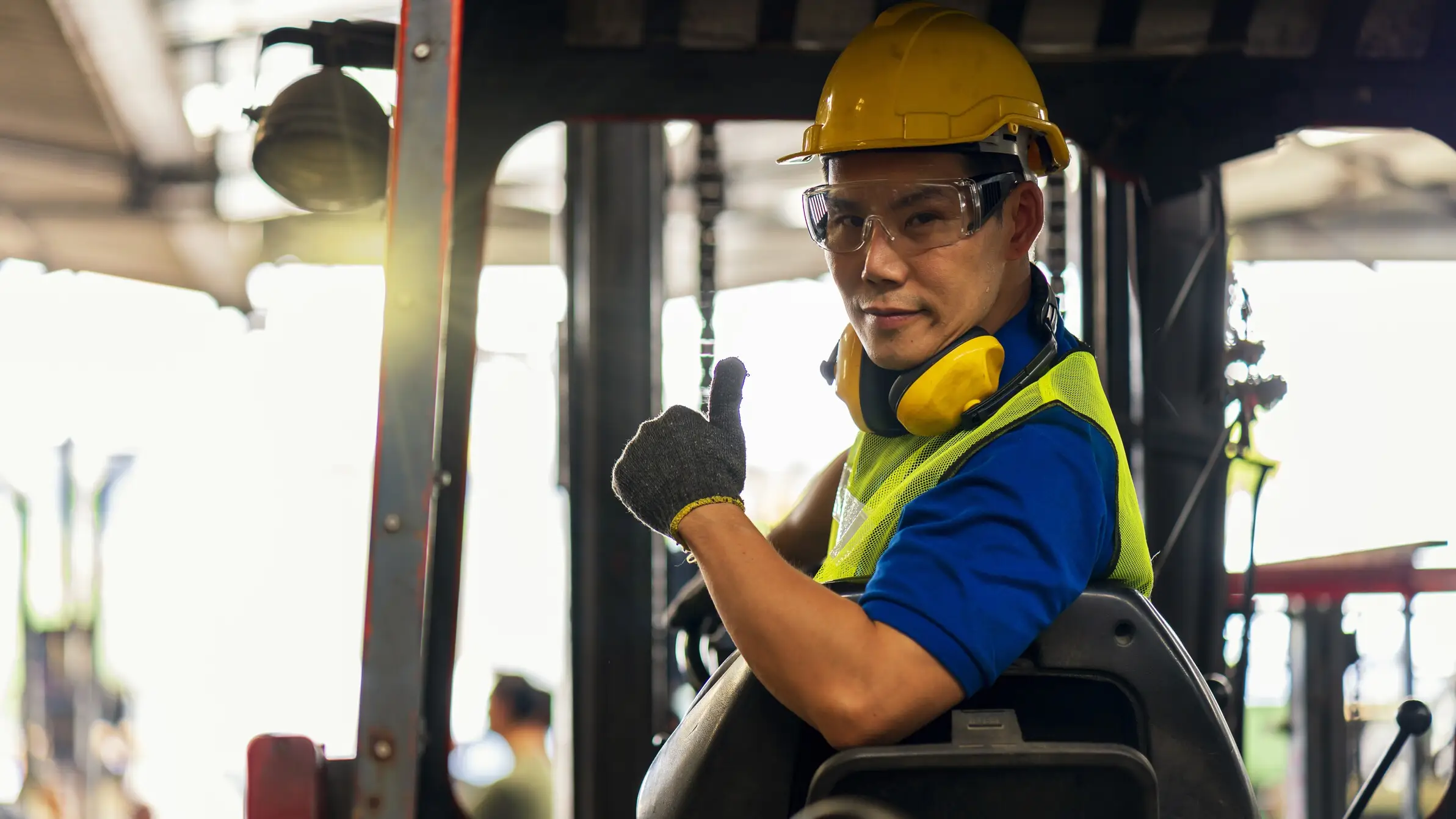 Forklift operator in a hard hat, safety glasses and hi-vis vest giving a thumbs up from the cab of a forklift in a warehouse loading area.