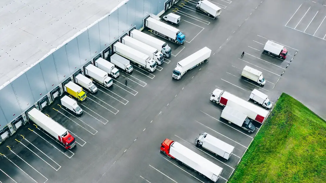 Aerial view of an industrial loading bay with multiple trailers docked at bays alongside a warehouse, and additional trucks and vans parked in the yard.