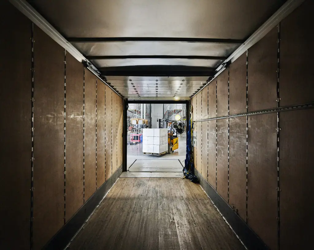 Wide view from inside of truck trailer of warehouse worker using forklift to load pallet of goods