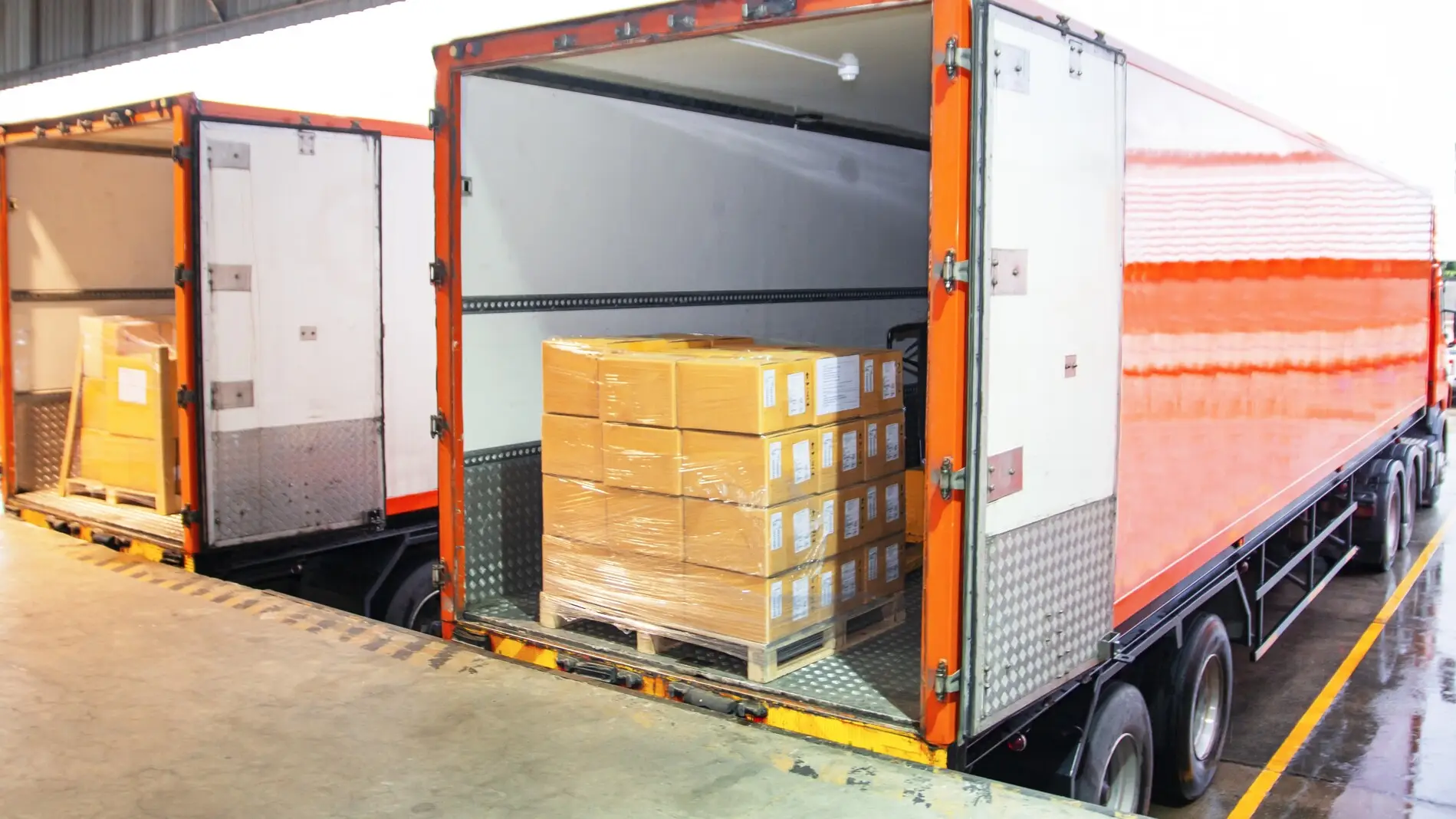 Open trailer backed up to a loading bay dock, with pallets of shrink-wrapped boxes ready to be loaded, and a safety barrier net across the open trailer doors.