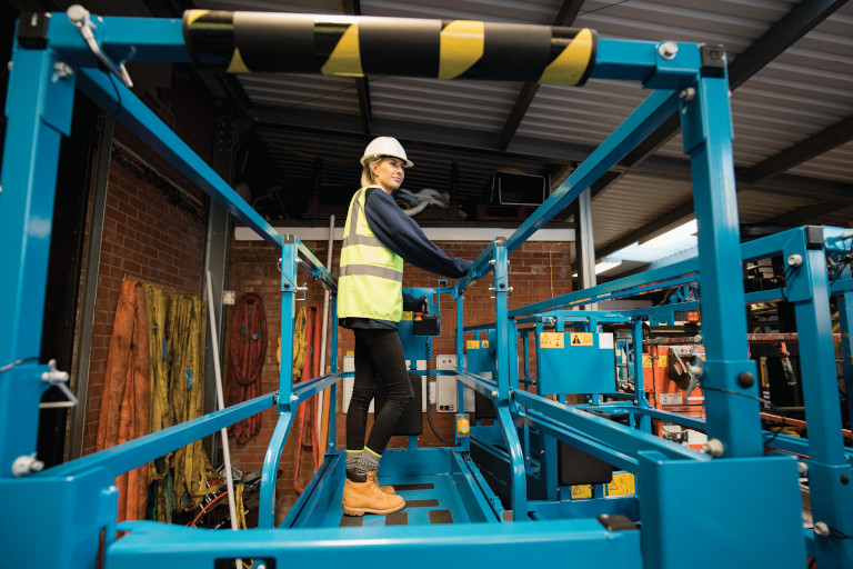A low-down shot of a trainee mechanic standing on a cherry picker, she is wearing a protective hard hat, hi-vis jacket and warm clothing.